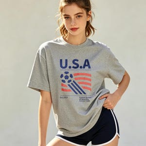 Op de afbeelding: Grijs T-shirt met de tekst "U.S.A" in marineblauw, een voetbal en een rood-blauw streepontwerp. Het shirt bevat ook de tekst "Est. 1913" en "United States of America Soccer Club".