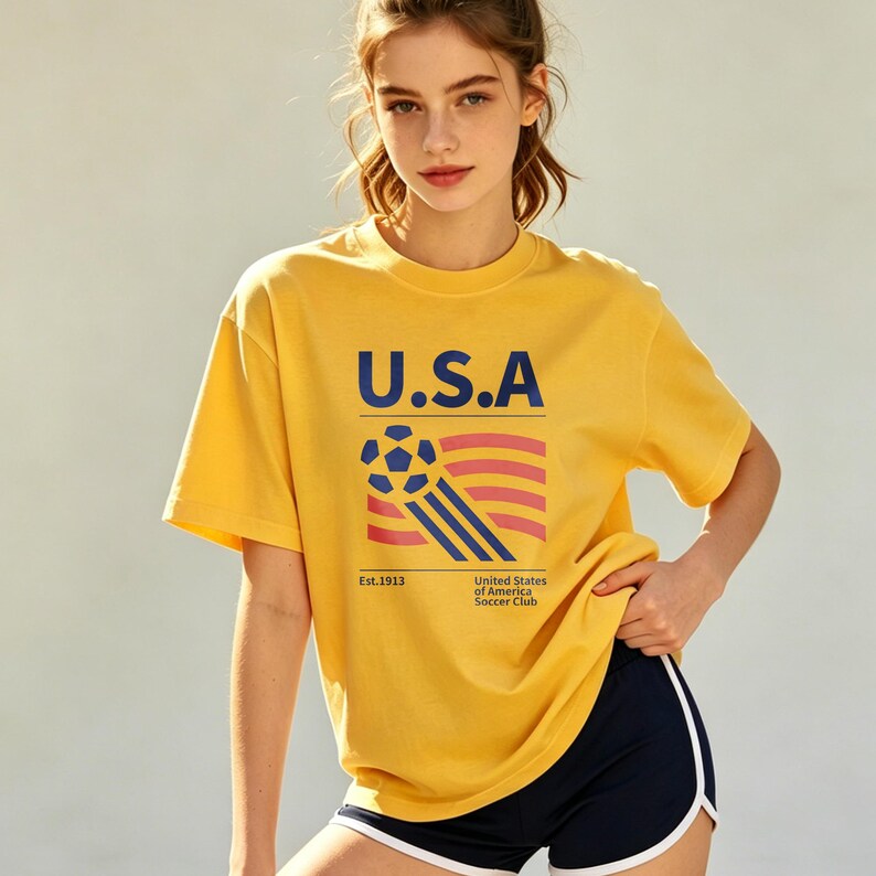 Op de afbeelding: Mosterdgeel T-shirt met de tekst "U.S.A" in marineblauw, samen met een voetbal en een vlagafbeelding. Het shirt bevat ook de tekst "Est. 1913" en "United States of America Soccer Club".