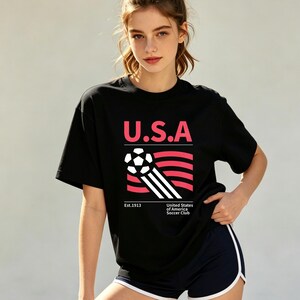 Op de afbeelding: Zwart T-shirt met de tekst "U.S.A" in roze, boven een voetbalafbeelding en rood-witte strepen. Het shirt bevat ook de tekst "Est.1913" en "United States of America Soccer Club".