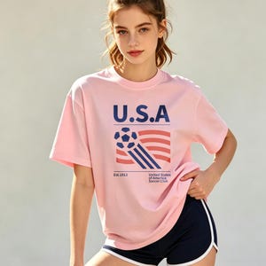 Op de afbeelding: Lichtroze T-shirt met de tekst "U.S.A" in marineblauw, samen met een voetbal en een gestreept ontwerp. Het shirt bevat ook de tekst "Est.1913" en "United States of America Soccer Club".