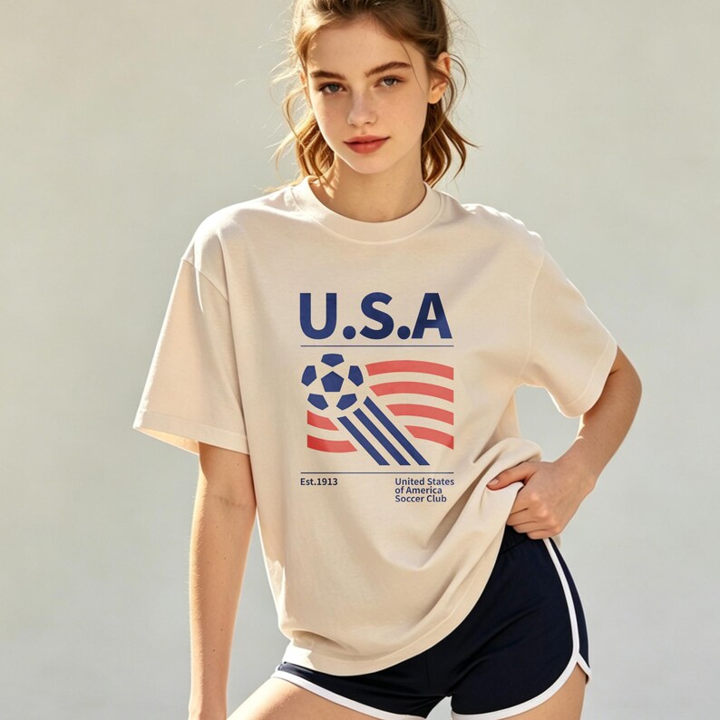 Op de afbeelding: Cr&egrave;mekleurig T-shirt met de tekst "U.S.A" in marineblauw, samen met een voetbal en een vlagafbeelding. Het shirt heeft ook de tekst "Est.1913" en "United States of America Soccer Club". De persoon draagt een marineblauwe short met witte bies.