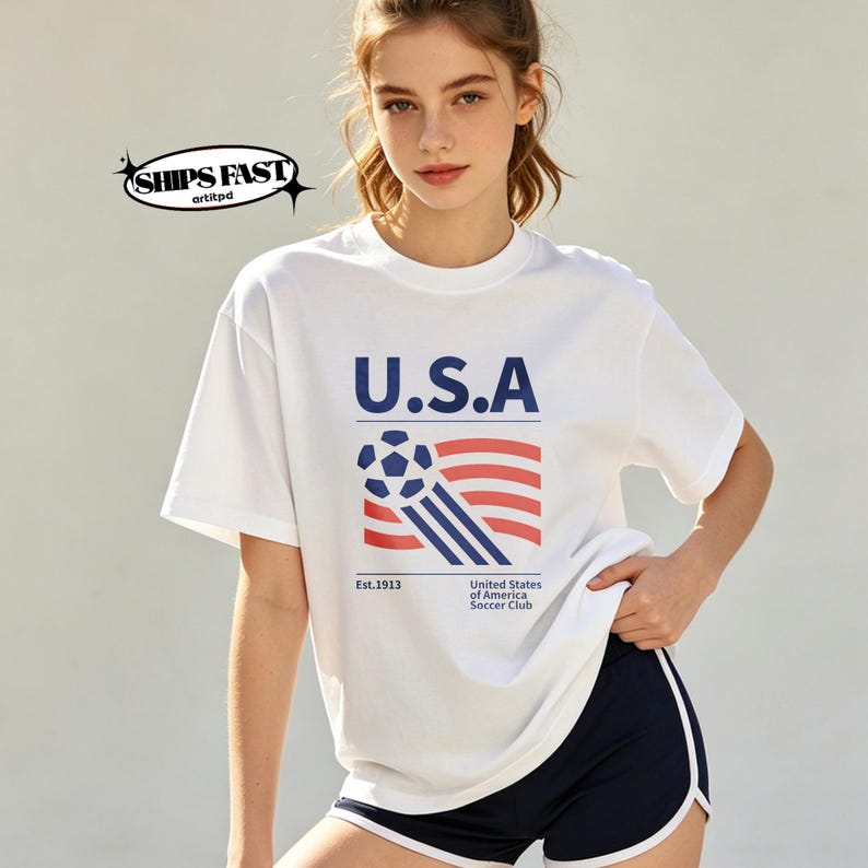 Op de afbeelding: Wit T-shirt met de tekst "U.S.A." in marineblauw, een voetbal en een gestileerd Amerikaans vlagontwerp. Het shirt bevat ook de tekst "Est.1913" en "United States of America Soccer Club".