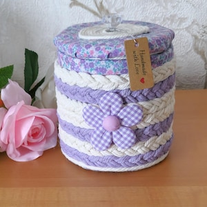 Puede incluir: Un recipiente de almacenamiento cilíndrico envuelto en filas alternas de cordón trenzado blanco y lavanda. La tapa presenta un patrón de tela floral y un pomo transparente. Una flor de tela con un patrón de cuadros vichy lavanda está unida al lateral.