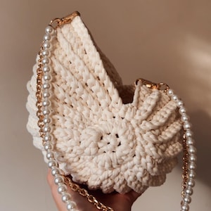 May include: Cream-colored, crocheted handbag with a pearl and gold chain strap. The bag has a textured, woven design and gold-toned hardware. The pearl strap adds a touch of elegance to this handmade accessory.
