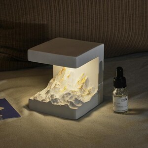 May include: A white, cube-shaped lamp with a mountain range design on the front. The mountain range has gold accents and is illuminated from within. A small glass bottle with a black dropper is next to the lamp.