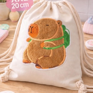 Könnte beinhalten: Cremefarbener Kordelzugbeutel mit einem braunen, orangefarbenen und grünen Capybara-Design. Der Beutel ist mit "XXL 20 cm" beschriftet. Das Capybara hat einen grünen Rucksack und ist mit Pailletten bestickt. Der Beutel hat einen Kordelzugverschluss.
