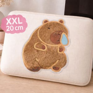 Könnte beinhalten: Eine beige Laptop-Hülle mit einer braunen Bärenillustration und einem blauen Tränentropfen. Der Bär ist mit dekorativen Stichen umrandet. Die Hülle ist mit "XXL" gekennzeichnet und 20 cm groß.