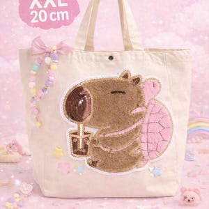 Könnte beinhalten: Beige Tragetasche mit einem Capybara-Design. Das Capybara ist braun mit einem rosa Panzer und hält ein Getränk. Die Tasche hat eine rosa Schleife und den Text "XXL 20 cm".