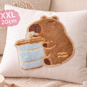 Könnte beinhalten: Ein weißes quadratisches Kissen mit einem braunen Capybara-Design, das ein blaues und braunes Getränk hält. Das Kissen hat ein rosa Etikett mit der Aufschrift "XXL 20cm". Das Capybara ist braun mit goldenen Pailletten-Akzenten. Das Getränk ist hellblau und braun.