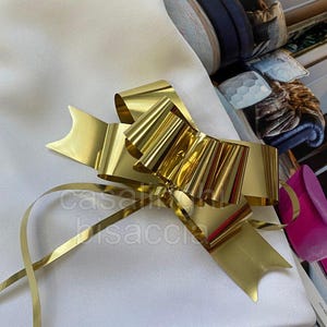 Puede incluir: Un lazo dorado brillante con largas cintas, sobre una tela blanca. El lazo tiene un diseño en capas con un aspecto texturizado. El texto "casasacchi" es parcialmente visible en la cinta.