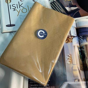 Op de afbeelding: Een rechthoekig pakket verpakt in bruin papier en verzegeld met een transparante plastic verpakking. Een ronde sticker met de tekst "ASALINGHI" is op de voorkant bevestigd. Het pakket ligt op een wit oppervlak naast een tijdschrift.