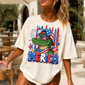 May include: An off-white t-shirt featuring a graphic of a green alligator wearing a baseball cap and star-shaped sunglasses, both with American flag patterns. The design includes fireworks, stars, and the text MERICA in red, white, and blue.