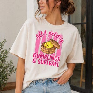 May include: Cream-colored t-shirt with a graphic that reads "All I need is Dumplings & Softball." The graphic features a cartoon dumpling in a bamboo steamer, set against a pink striped background. The shirt is a casual, everyday wear item.