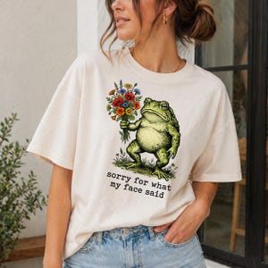 May include: A cream-coloured t-shirt with a detailed illustration of a green frog holding a bouquet of colourful flowers. The text "sorry for what my face said" is printed below the frog. A casual, everyday wear item.