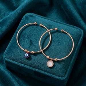 May include: Two gold-colored open cuff bracelets. One bracelet has a blue and white evil eye charm, and the other has a white circular charm with a crystal border. The bracelets are displayed on a teal velvet surface.