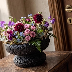 May include: A floral arrangement in a black, textured, two-tiered vase. The flowers include deep red dahlias, pink roses, purple sweet peas, and small blue blossoms. The vase sits on a wooden surface.