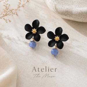 Könnte beinhalten: Ein Paar schwarze Blumenohrringe mit goldfarbenen Zentren und hellblauen Perlen. Der Hintergrund ist neutral, mit dem Text "Atelier The Muse" am unteren Rand.