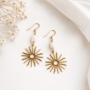 Könnte beinhalten: Ein Paar goldfarbene Ohrringe mit Sonnenmuster. Jeder Ohrring hat eine Süßwasserperle, die über dem Sonnenmuster hängt. Die Ohrringe sind vor einem weißen Hintergrund mit floralen Akzenten platziert.
