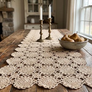 Op de afbeelding: Beige gehaakte tafelloper met geschulpte rand op een rustieke houten tafel. Twee messing kandelaars met witte kaarsen en een kom met peren staan ook op de tafel. De kamer heeft een gezellige, landelijke uitstraling.