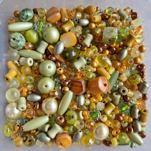 Op de afbeelding: Een verzameling diverse kralen in verschillende tinten groen, geel, amber en bruin. De mix bevat ronde, cilindrische, gefacetteerde en onregelmatige vormen van glas, plastic en hout, allemaal in een vierkante plastic bak.