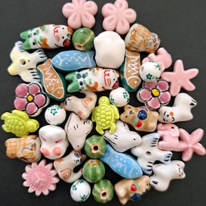 Op de afbeelding: Een verzameling diverse keramische kralen in verschillende vormen, waaronder bloemen, vissen, schildpadden, katten en ronde bollen. De kralen hebben een palet van roze, blauw, groen, wit en bruin, waarbij sommige stukken handgeschilderde details bevatten.