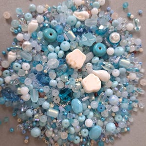 Op de afbeelding: Een verzameling losse knutselkralen in diverse tinten blauw, wit en turkoois. Het assortiment bevat ronde, gefacetteerde, cilindrische en stervormige kralen, samen met verschillende kleine witte schelpvormige bedels die erdoorheen gemengd zijn.