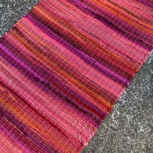 May include: A rectangular, handwoven rug with a vibrant color palette of pink, magenta, and orange hues. The rug features a striped pattern with a fringed edge. The texture appears to be made of woven fabric.