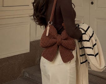 May include: A brown, bow-shaped crochet handbag with a long strap. The bag is a deep shade of brown and features a large bow design. The strap is also brown. The bag is worn over the shoulder.