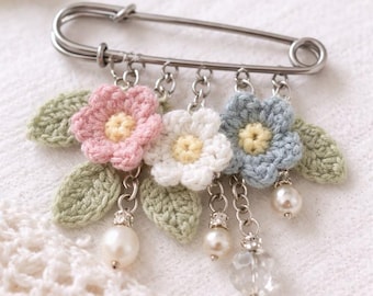 May include: A decorative safety pin featuring three crocheted flowers in pink, white, and blue, each with a yellow center. The flowers are accented with green leaves and dangling pearl and crystal charms. A charming accessory.