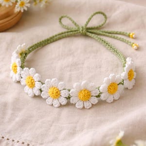 Può includere: Una fascia per capelli all'uncinetto fatta a mano con fiori di margherita bianchi con centri gialli. La fascia è collegata da una fascia verde all'uncinetto che si annoda con un fiocco sul retro. Le margherite sono uniformemente distanziate.