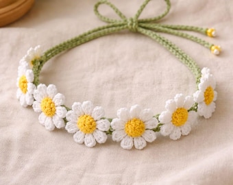 May include: A handmade crochet headband featuring white daisy flowers with yellow centers. The headband is connected by a green crocheted band that ties in a bow at the back. The daisies are evenly spaced around the band.