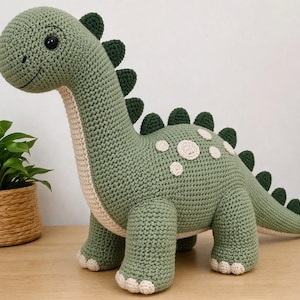 May include: A handmade, green crocheted dinosaur toy with a long neck, dark green spines, and cream-colored accents. The dinosaur has white circular spots on its back and a friendly expression. The toy is approximately 18 inches long.