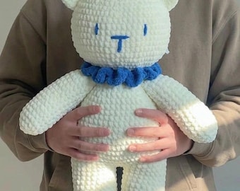 May include: A white crocheted teddy bear plush toy with a textured yarn finish. The bear features simple blue embroidered eyes and a blue nose, and it wears a ruffled blue yarn collar around its neck. The toy has rounded ears and limbs.