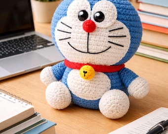 May include: A handmade blue and white crochet plush toy of a cartoon character with a red nose and collar, and a yellow bell. The toy is sitting on a wooden desk next to a laptop, books, and a pen.