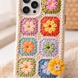 May include: A cream-colored crochet phone case featuring a colorful floral design. The case has a grid pattern of squares, each with a different colored flower in the center. The flowers are in shades of pink, orange, yellow, and blue. The case is designed to fit a smartphone.