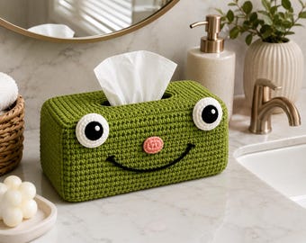 May include: A rectangular olive green crochet tissue box cover featuring two large white and black circular eyes, a small pink circular nose, and a black embroidered curved mouth, with a white tissue protruding from the top opening.