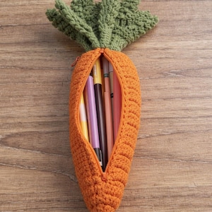 Può includere: Astuccio per matite all'uncinetto arancione fatto a mano a forma di carota, con foglie verdi in cima. L'astuccio è aperto, rivelando diverse penne e matite colorate. Posizionato su una superficie di legno.