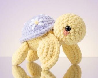 May include: A crocheted turtle plush toy with a pale yellow body and a light purple shell. The shell features a white daisy flower with a yellow center. The turtle has black plastic eyes and small pink stitched cheeks on its round head.