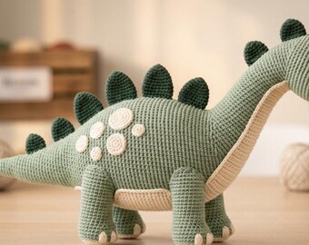 May include: A sage green crochet dinosaur toy featuring a long neck, a cream-colored underbelly, and dark green triangular plates along its back. The dinosaur has small cream-colored circular patches on its side and black plastic safety eyes.