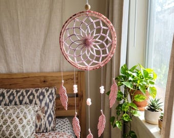 May include: A pink dreamcatcher with a circular frame and intricate web design. Pink feathers and beads hang from the bottom. The dreamcatcher is suspended in front of a bed with patterned pillows and a wooden headboard.