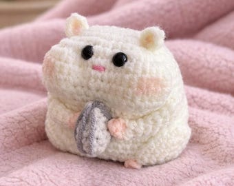May include: A handmade, white crochet hamster with black eyes, pink cheeks, and a gray sunflower seed. The plushie is sitting on a pink, textured surface. The hamster is holding a gray sunflower seed.