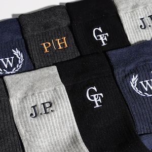 Könnte beinhalten: Eine Auswahl an gerippten Socken in verschiedenen Farben, darunter Marineblau, Schwarz und Grau. Jede Socke zeigt gestickte Initialen oder ein Monogramm-Design in Kontrastfarben wie Gold und Weiß. Die Socken sind ordentlich angeordnet.