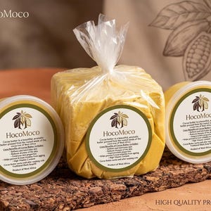 Könnte beinhalten: Ein Bild von Kakaobutterprodukten, darunter ein verpackter Block und zwei runde Behälter. Die Produkte tragen Etiketten mit dem Markennamen "HocoMoco" und Produktinformationen. Die Kakaobutter hat eine hellgelbe Farbe.