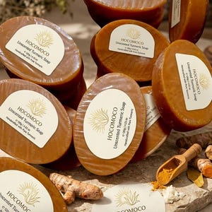 May include: Oval-shaped, brown unscented turmeric soaps are stacked, some with labels reading "HocoMoco Unscented Turmeric Soap." The image includes turmeric root, powder, and a wooden scoop. The text "high quality product" is at the bottom.