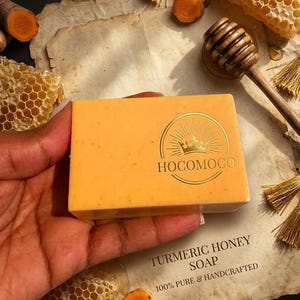 May include: A rectangular turmeric honey soap bar with a gold logo, held in a hand. The soap is surrounded by turmeric slices, honeycomb, and a wooden honey dipper. Text on the packaging reads "Turmeric Honey Soap" and "100% Pure & Handcrafted."