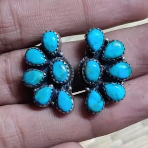 May include: A pair of silver-toned stud earrings, each featuring a cluster of turquoise-colored stones arranged in a floral pattern. The earrings are held in a hand against a neutral background.