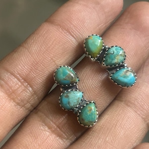 Może przedstawiać: Para turkusowo-srebrnych kolczyków. Każdy kolczyk ma trzy turkusowe kamienie w kształcie łezki osadzone w srebrnej oprawie. Turkusowe kamienie mają nakrapiany wygląd z odcieniami niebieskiego i zielonego.