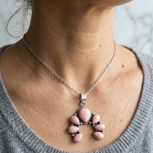 May include: A silver-toned necklace featuring a crescent-shaped pendant adorned with seven smooth, oval-shaped pink stones. The pendant is suspended from a delicate silver-toned chain, showcasing a symmetrical arrangement of the pink cabochons.