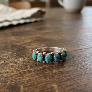 Puede incluir: Un anillo de plata con una hilera de cinco piedras de turquesa de forma ovalada engastadas en biseles individuales. La banda está decorada con pequeñas cuentas de plata situadas entre cada piedra, creando un patrón decorativo continuo.