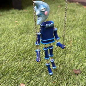 May include: A marionette puppet featuring a light blue crescent moon head with pink cheeks, a dark blue cylindrical torso with a small bow tie, and jointed dark blue limbs. The figure is suspended by silver metal chains attached to its head and arms.